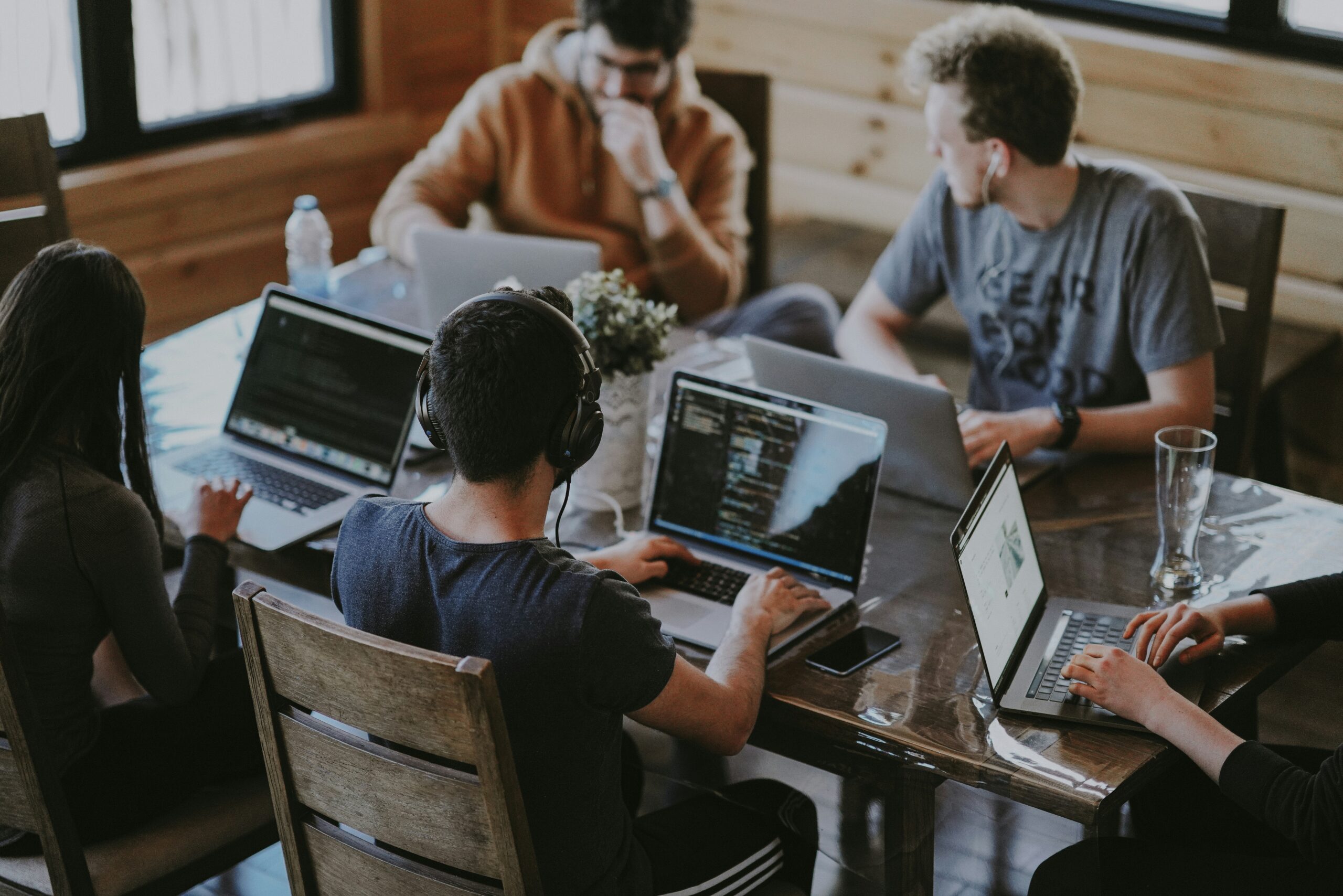 Team of professionals collaborating around a wooden table with laptops in a modern office setting, engaged in discussion and teamwork