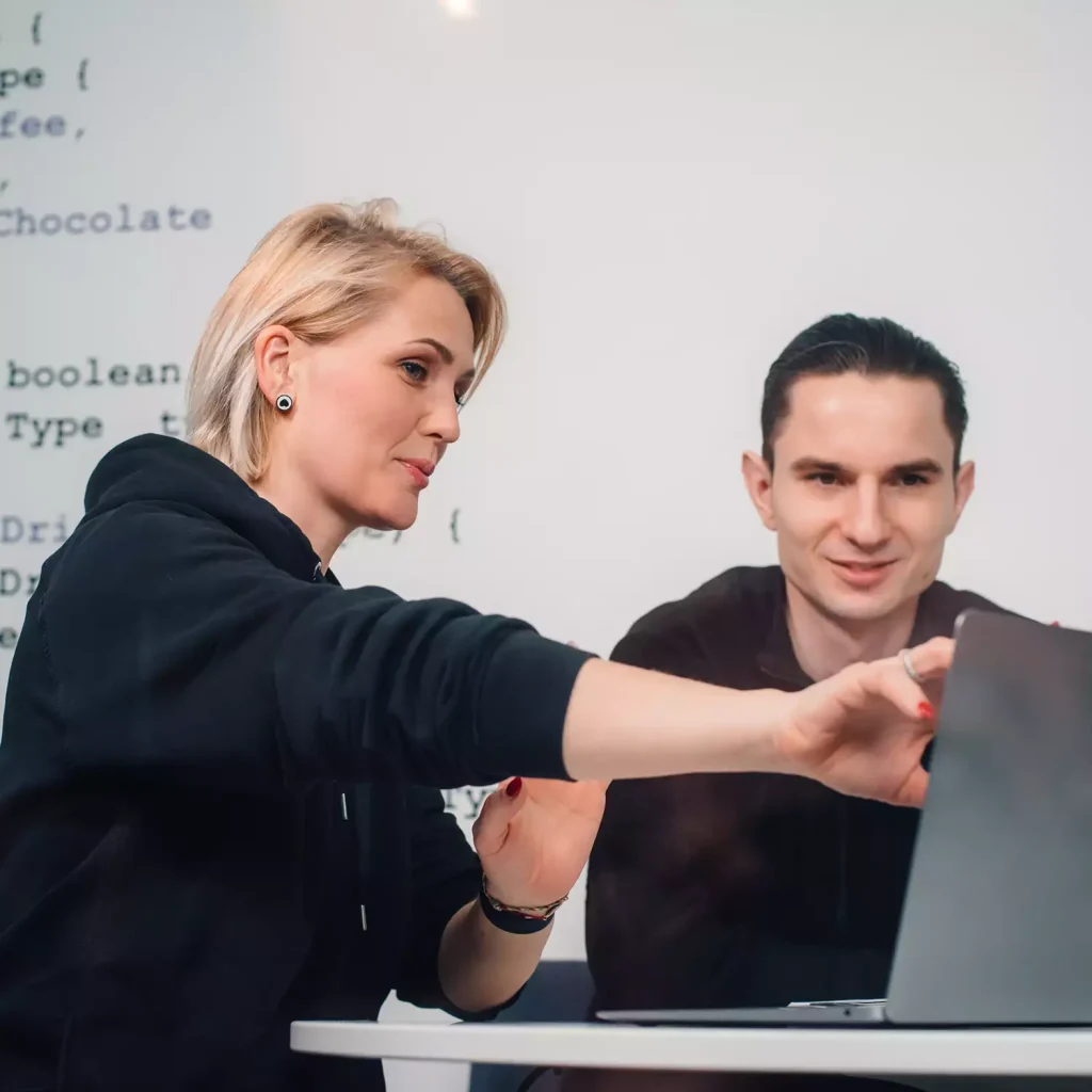 Two Softeta colleagues collaborating at a laptop, with code displayed on a screen in the background